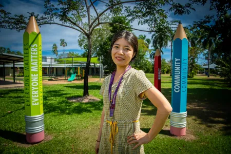 A young Chinese woman stands outside surrounded by larger than life pencils in vibrant colours. The pencils are as tall as she is and have words on them like 'everyone matters,' 'community,' and 'respect.'